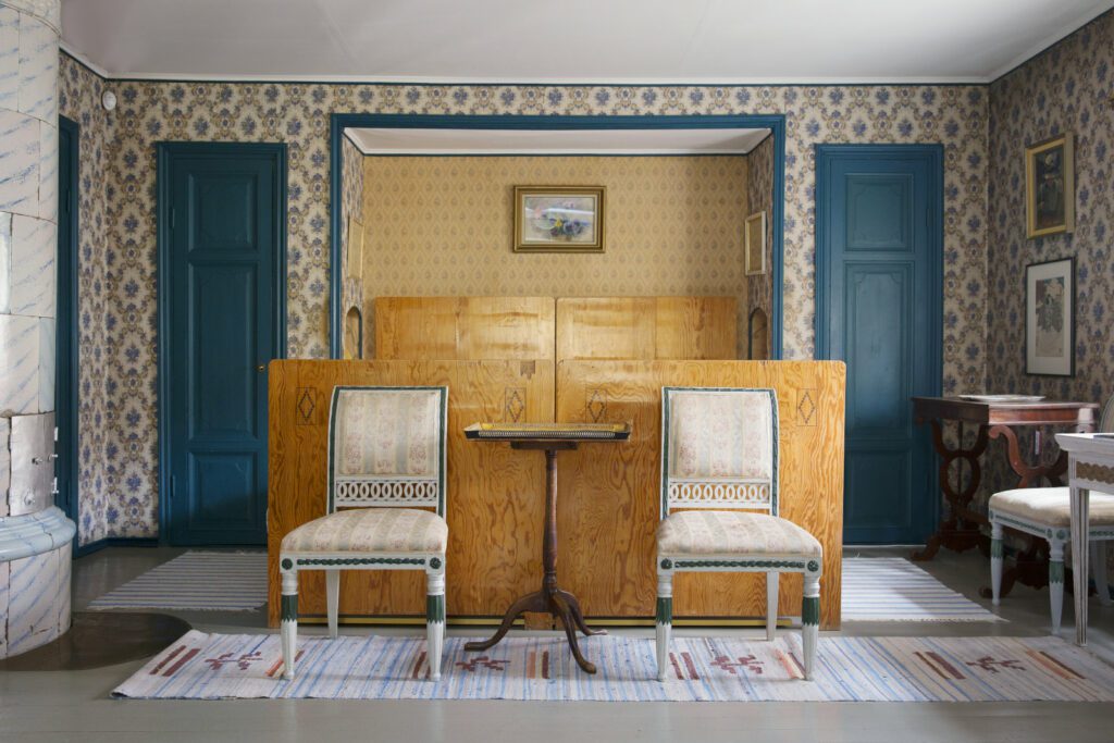 Casa Haartmannin makuuhuone, vaaleanruskea sängynpääty, jota vasten kaksi vaaleaa vanhanaikaista tuolia. Kukkatapettinen seinä ja sinertävät ovet.