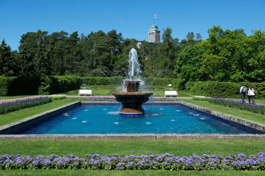 Suihkulähde ja sininen allas. Ympärillä Kultarannan puutarha ja kaksi matkailijaa. Taustalla Kultarannan kivilinna, jonka tornissa liehuu Suomen lippu.