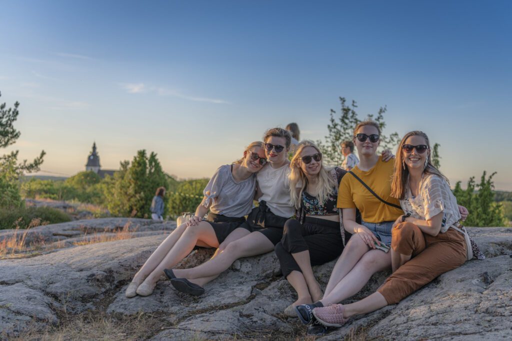Viisi aurinkolasipäistä tyttöä istuu Kuparivuoren kallioilla kesällä, taustalla Naantalin kirkon siluetti.