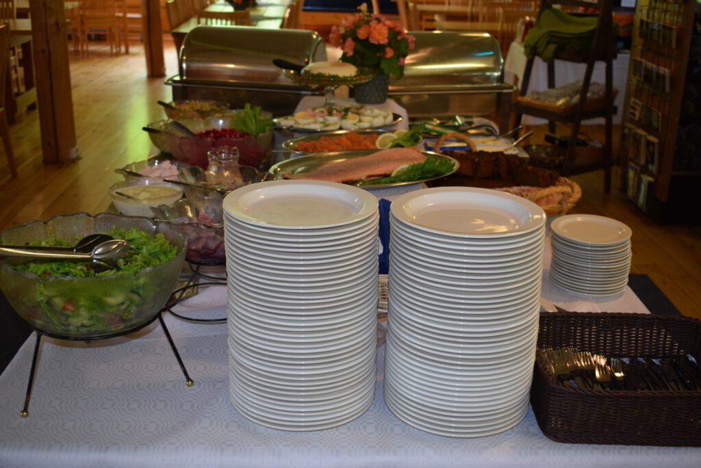 Buffetpöydän pää, kaksi korkeaa pinoa lautasia. Taustalla näkyy tarjoiluastioissa salaattia, lohta, rosollia, kananmunia ja leipää.
