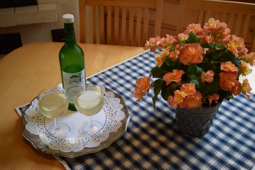 Pöydällä sinivalkoisen ruutuliinan päällä tarjottimella kaksi lasillista Louhisaaren juomaa ja vihreä pullo. Pöydällä ruukussa oransseja kukkia.
