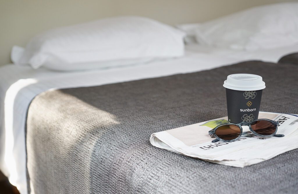 Naantali City Apartmentsin sänky, jonka päiväpeiton päällä sanomalehti, take away -kahvikuppi ja aurinkolasit.