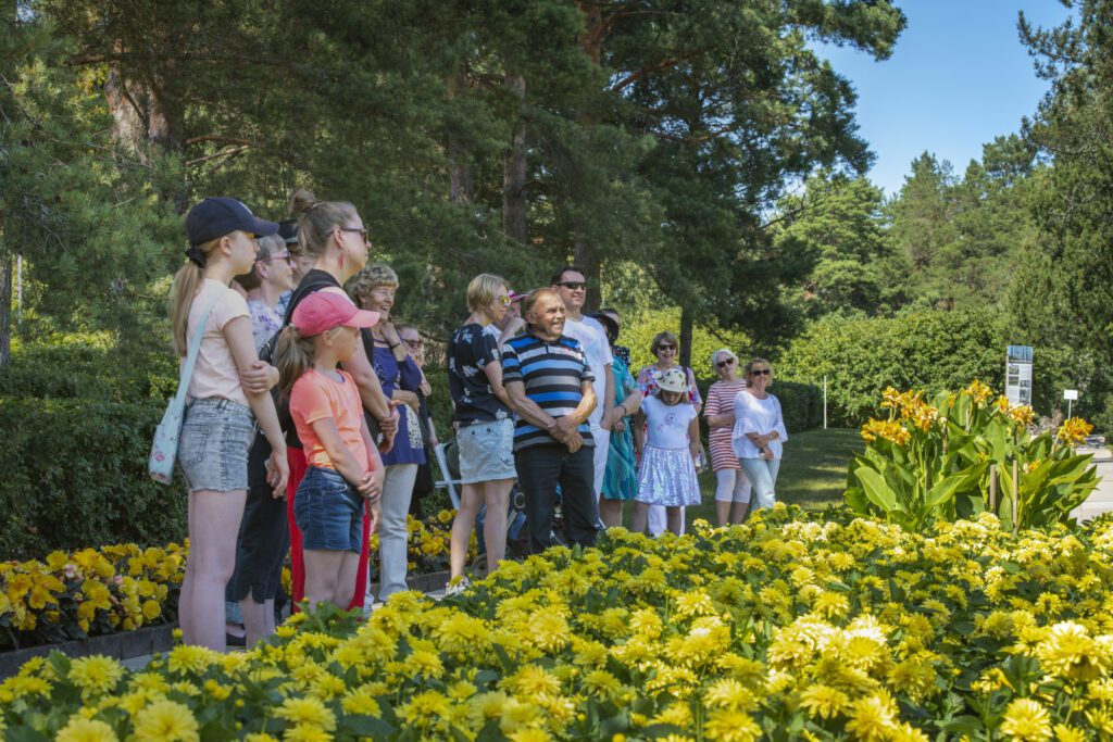 Joukko Kultarannnan puutarhan opastukselle osallistuvia asiakkaita. Keltaisia kukkia etualalla, puiston puita taustalla. Aurinkoinen päivä ja iloinen tunnelma.