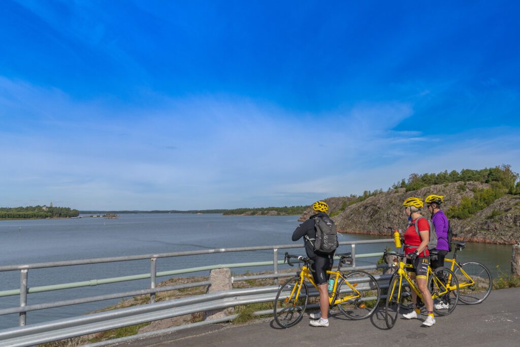 Kolme pyöräilijää katsoo merimaisemaa sillalta.