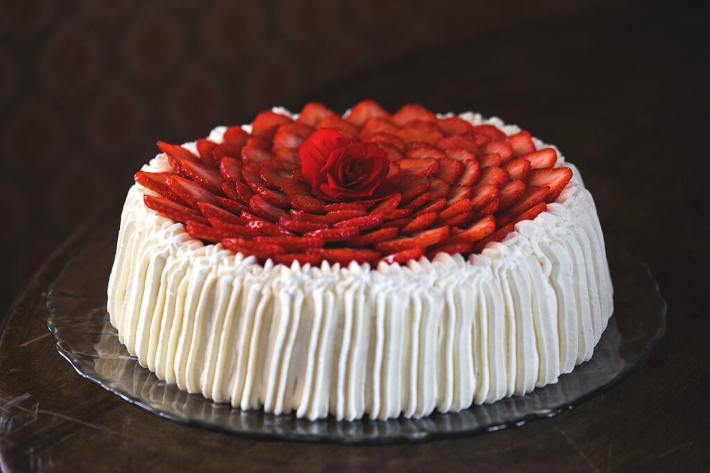Ravintola Pohjakulman täytekakku, valkoista kermaa reunoilla ja kakun päällä viipaloituja mansikoita ja punainen ruusu.