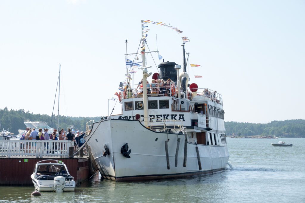 Valkoinen höyrylaiva Ukkopekka Naantalin laiturissa, lippuja liehuu kannella ja ihmisiä on sekä laivalla että laiturilla.