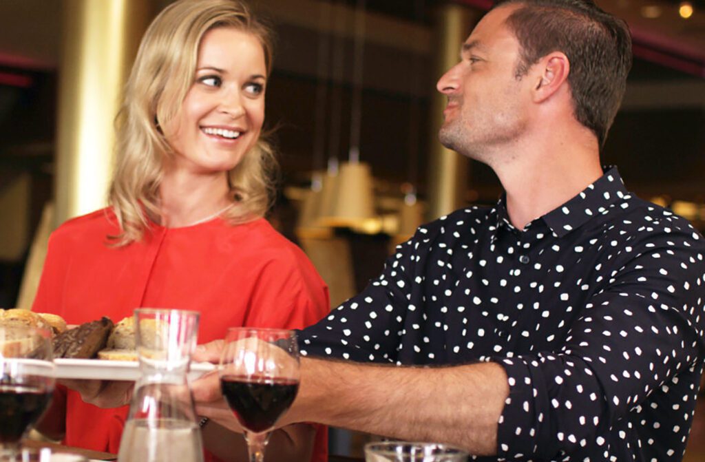 Punapaitainen nainen ja mustavalkopaitainen mies katsovat toisiaan ravintolassa ja hymyilevät toisilleen.