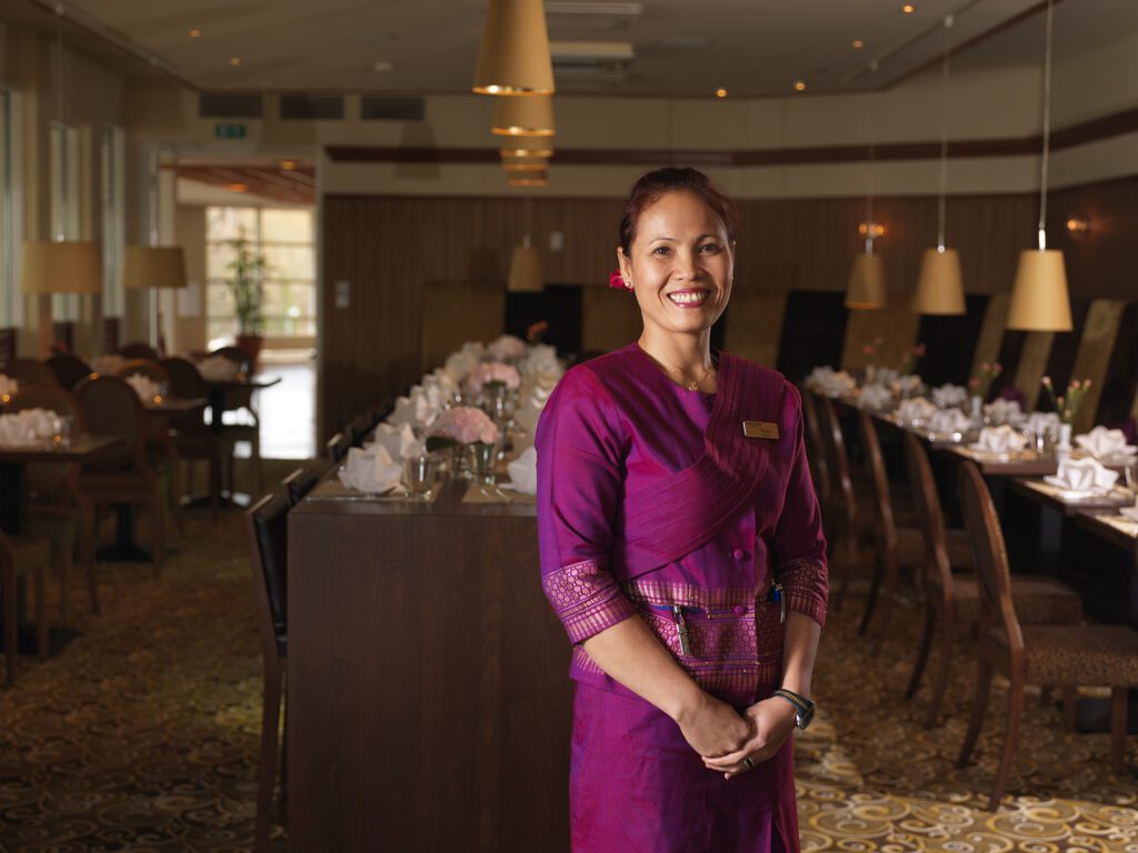 Purppuraan thaimaalaiseen asuun pukeutunut tarjoilija hymyilee Thai Garden -ravintolassa.