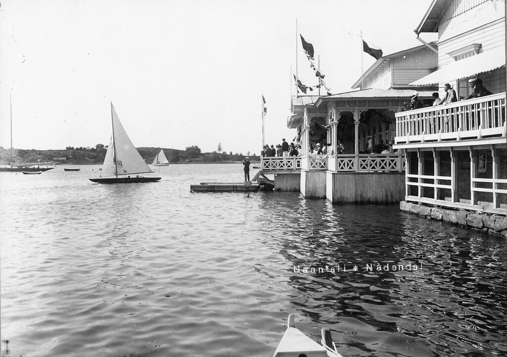 Mustavalkokuva Naantalin rannasta 1900-luvun alkupuolelta, ihmisiä katselemassa veneitä rannan rakennusten terasseilta.