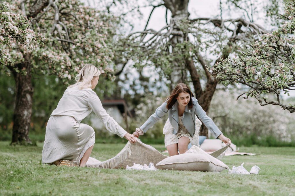 Kaksi nuorta naista levittämässä vilttiä omenapuiden alle puutarhaan.