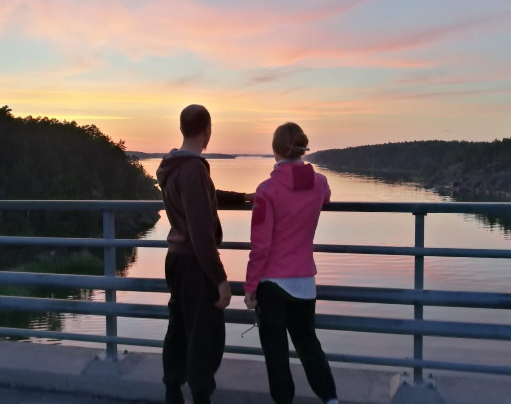 Nuori mies ja nainen katselevat sillalta alas merelle ja saaristoon hämärtyvässä kesäillassa.