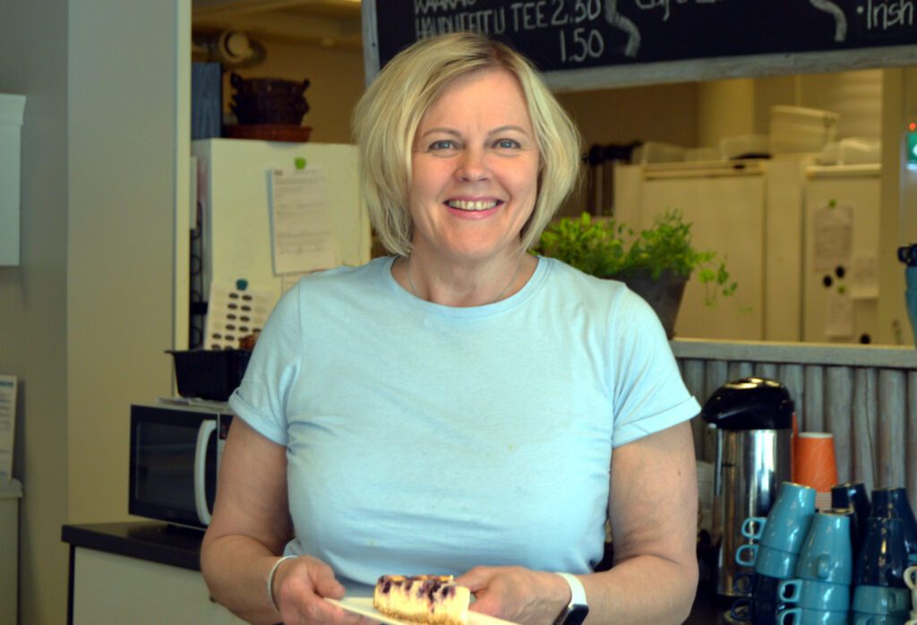Hymyilevä Cafe Akselin tarjoilija pitää kädessään lautasta, jossa mustikkakakun pala.