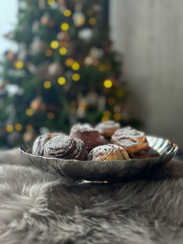 Ruokakummi-Annen piparinmakuiset pullakierteet kauniisti aseteltuna hopeiselle tarjoiluastialla, taustalla valaistu joulukuusi.