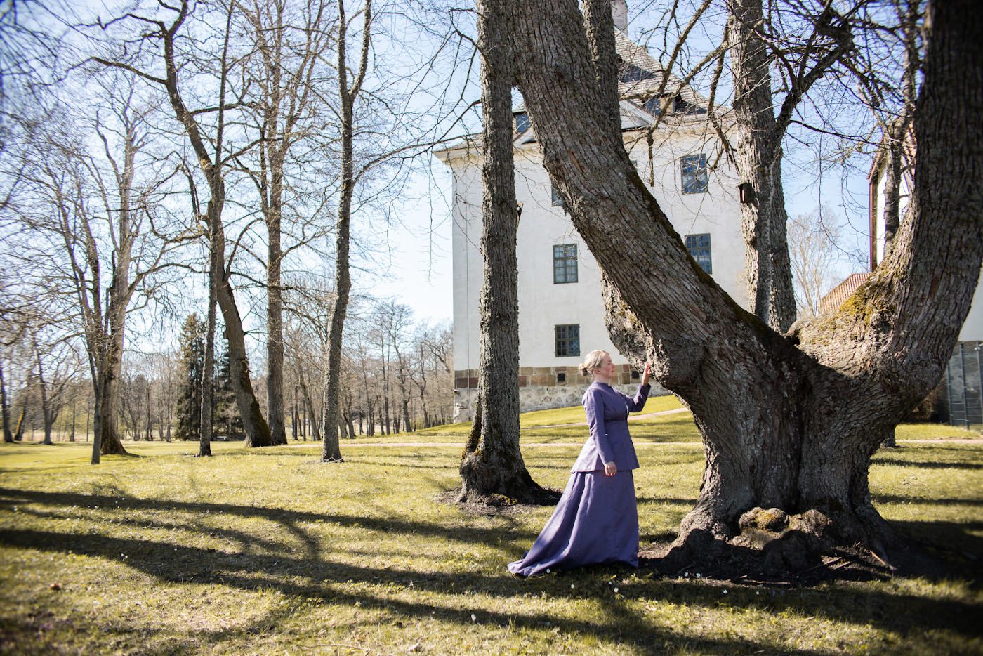 Vanhahtavaan, violettiin asuun pukeutunut nainen koskettaa vanhaa puuta Louhisaaren kartanolinnan puutarhassa.