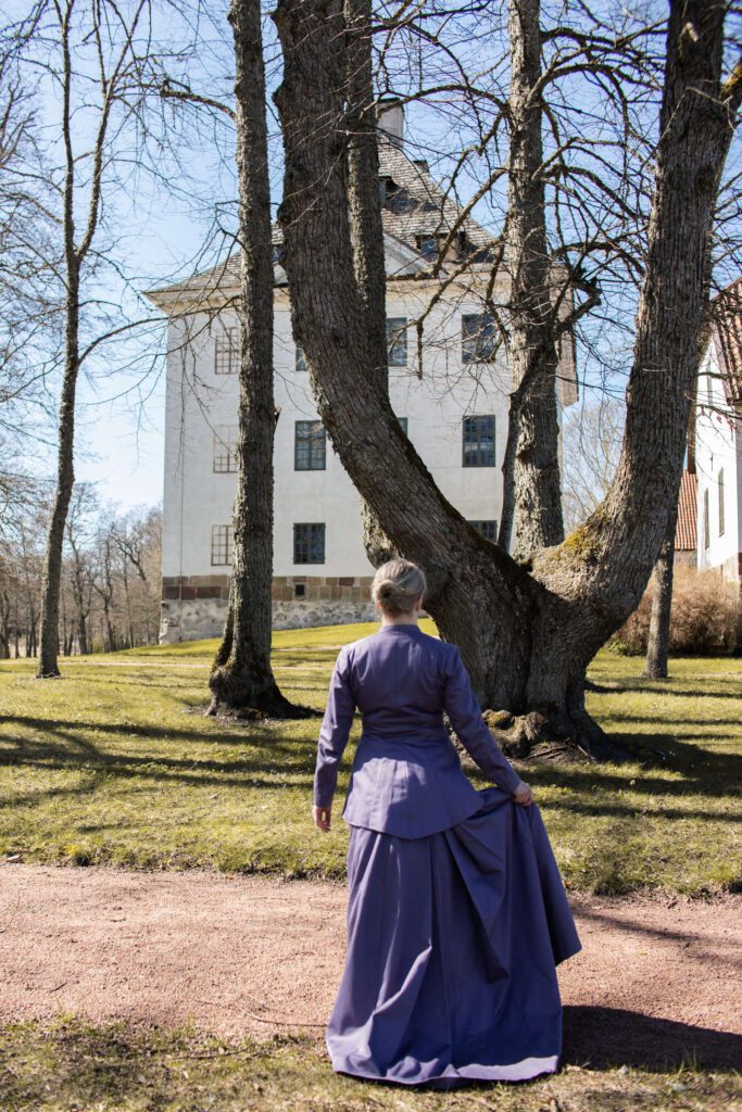 Vanhahtavaan asuun pukeutunut nainen kävelee Louhisaaren kartanolinnan puutarhassa kohti linnaa.