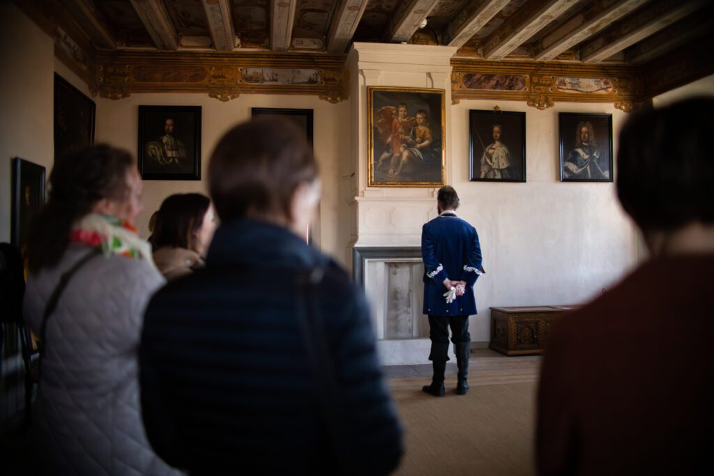 Näytelmäkierroksen miesnäyttelijä seisoo selin yleisöön ja katsoo seinällä olevia vanhoja maalauksia.