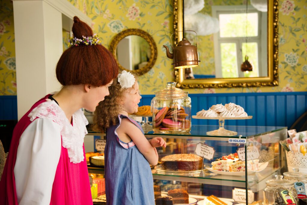 Mymmeli ja pieni tyttö katsovat vitriinin herkkuja kahvilassa, jossa on keltaiset seinät.