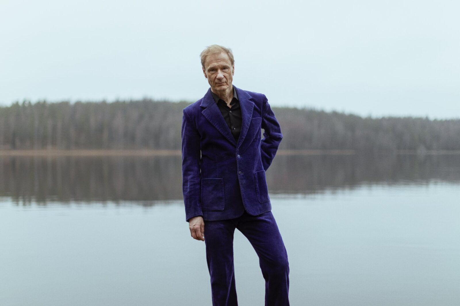Laulaja Ismo Alanko seisoo tummanlila puku päällään harmaa saaristomaisema taustanaan.