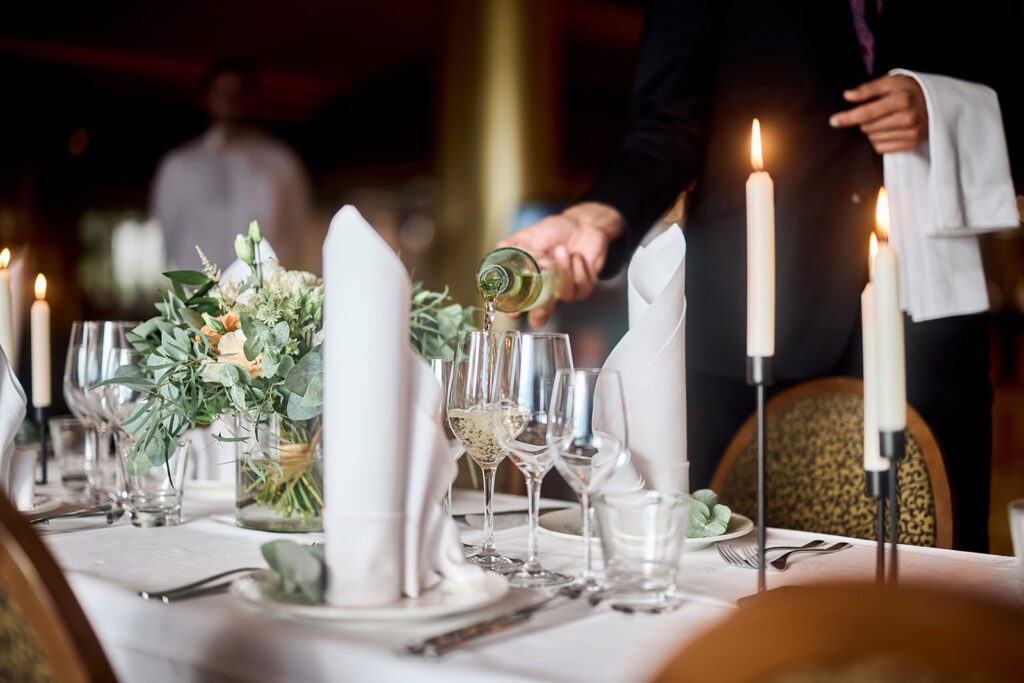 Juhlavasti katettu illallispöytä kynttilöiden loisteessa, tarjoilija kaataa viinilaseihin juotavaa.