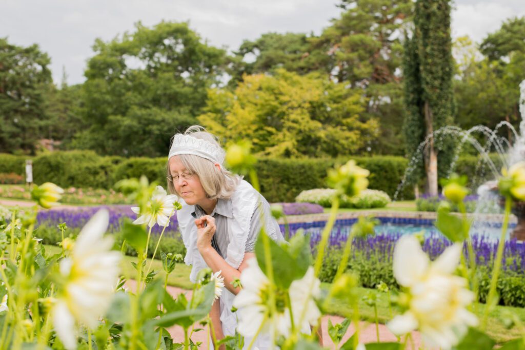 Mamselli-opas, jolla valkoinen hilkka ja essu, haistelee nautiskellen valkoista kukkaa, taustalla sinisiä kukkaistutuksia, pensasaitaa ja suihkulähde.
