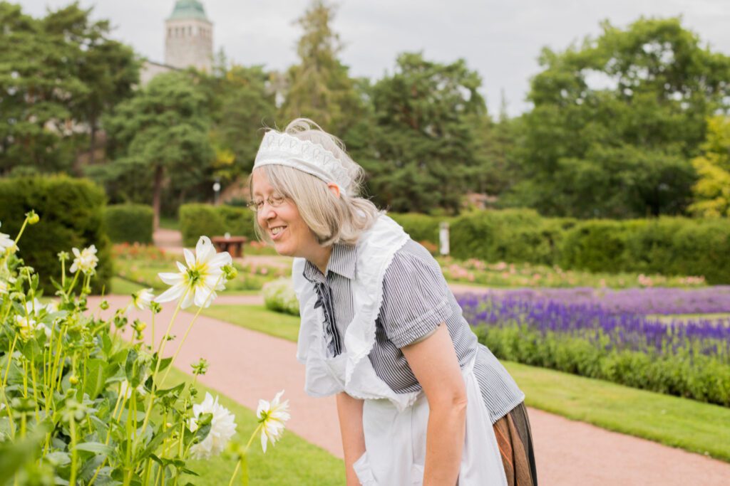 Hymyilevä Mamselli-opas haistelee isoa valkoista kukkaa Kultarannan puutarhassa.