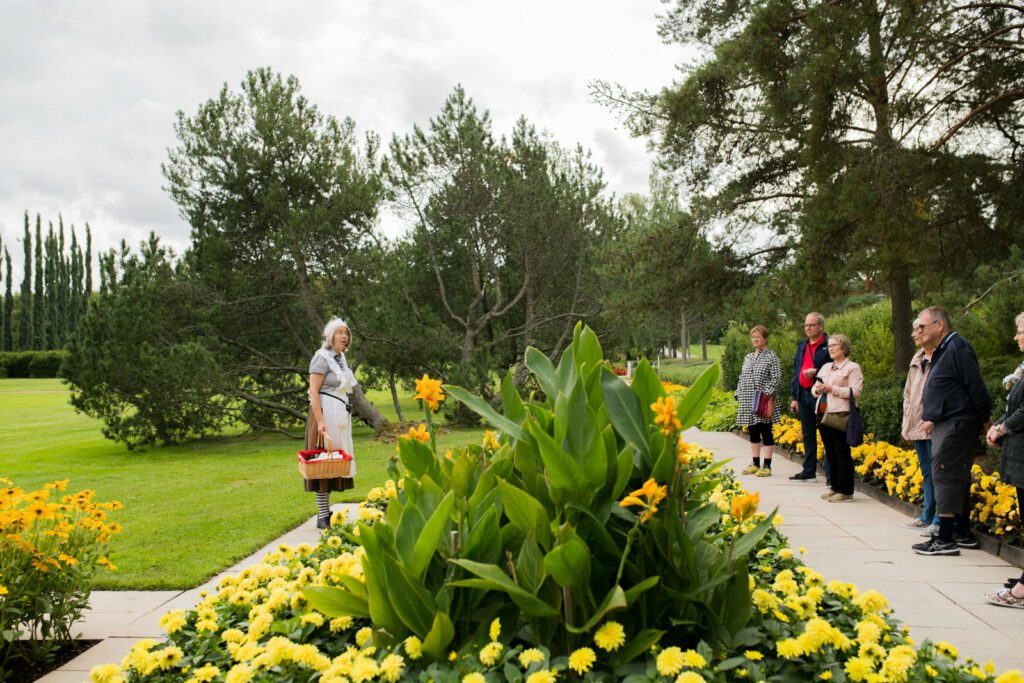 Harmaaseen mekkoon ja valkoiseen mekkoon pukeutunut Mamselli-opas esittelee keltaisia kukkia joukolle opastukselle osallistuvia matkailijoita.