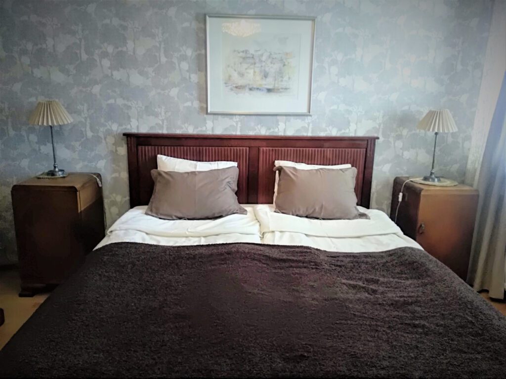Makuuhuoneen parivuode, ruskea päiväpeitto ja valkoiset lakanat, harmaa seinä ja maisemataulu.
