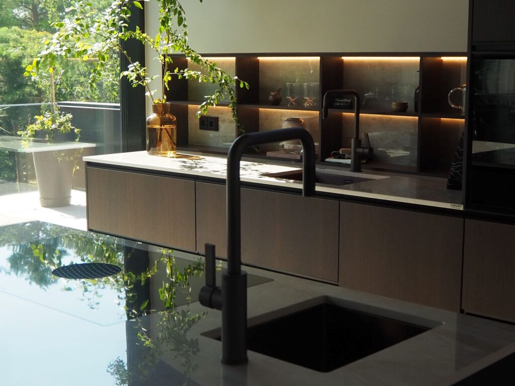 Tumman ruskea keittiö mustilla yksityiskohdilla ja iso ikkuna, josta tulvii tilaan valoa sekä luonnon vehreyttä.