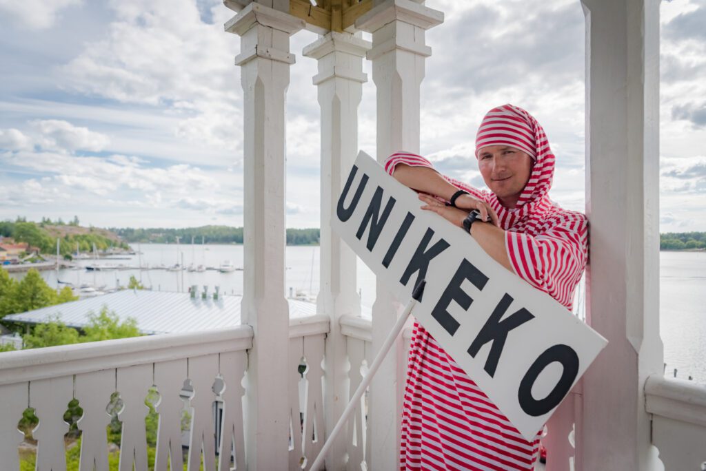 Mies Unikeko-puvussa nojailee näkötornin kaiteeseen Unikeko-kyltin kanssa. Taustalla merenlahti.
