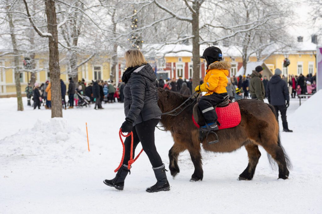 Nainen taluttamassa ponia, jonka selässä lapsi ratsastamassa Naantalin lumisessa Vanhassakaupungissa Lasten Joulu -tapahtumassa.