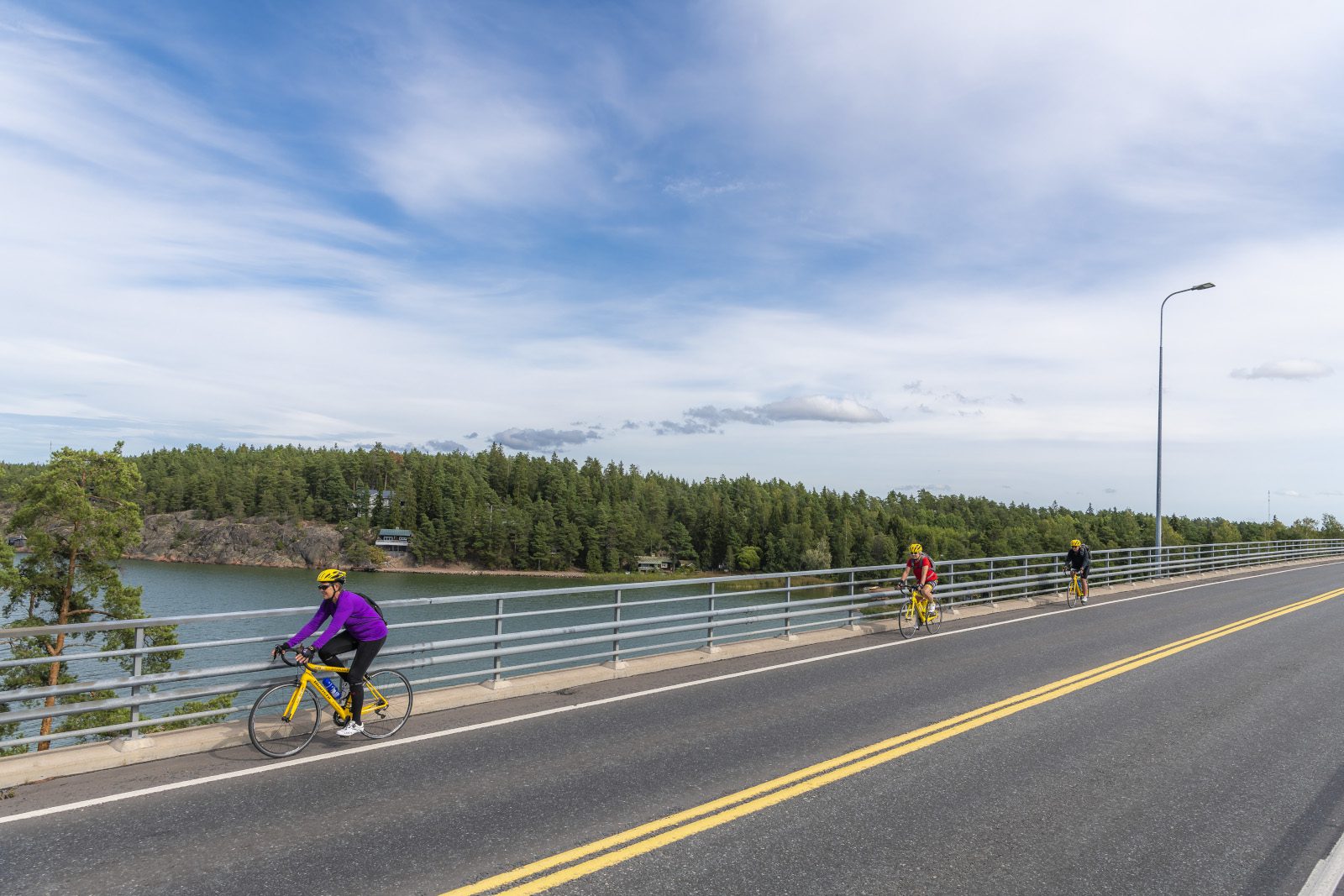 Kolme pyöräilijää pyöräilee sillan yli. Merta ja saarta näkyy sillan kaiteen takana.