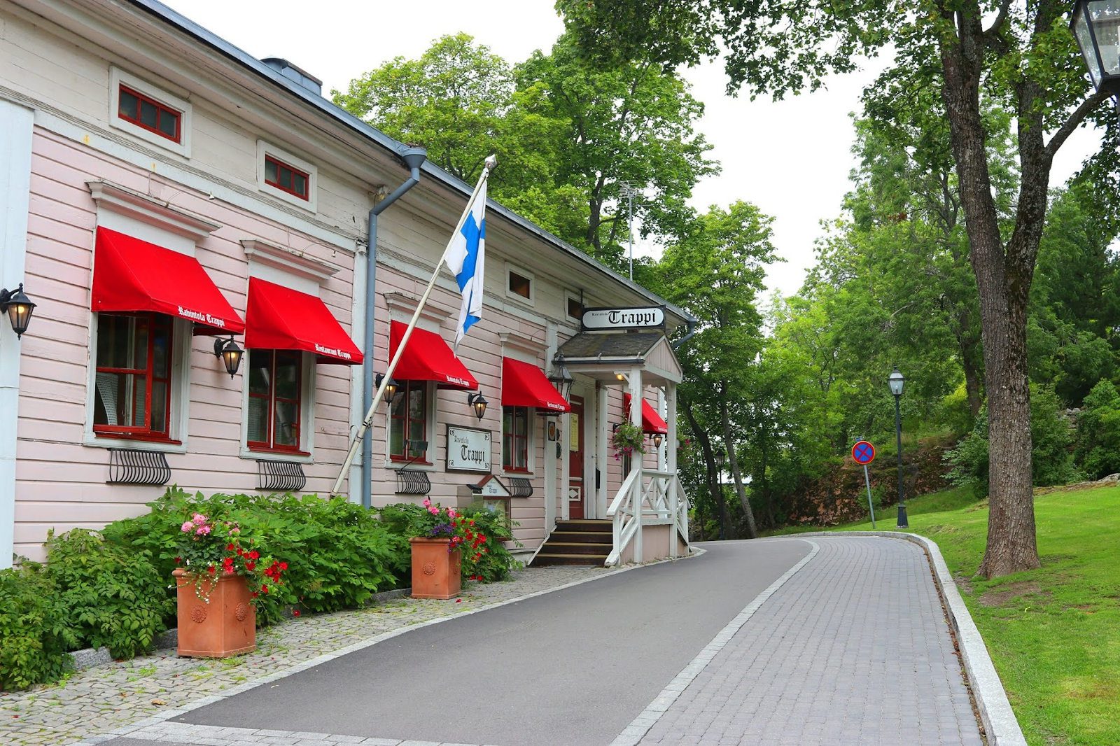 Vaaleanpunainen ravintolarakennus ja kivetty katu puiston varrella. Suomenlippu talon kyljessä, ikkunoissa punaiset markiisit, joissa lukee Ravintola Trappi.