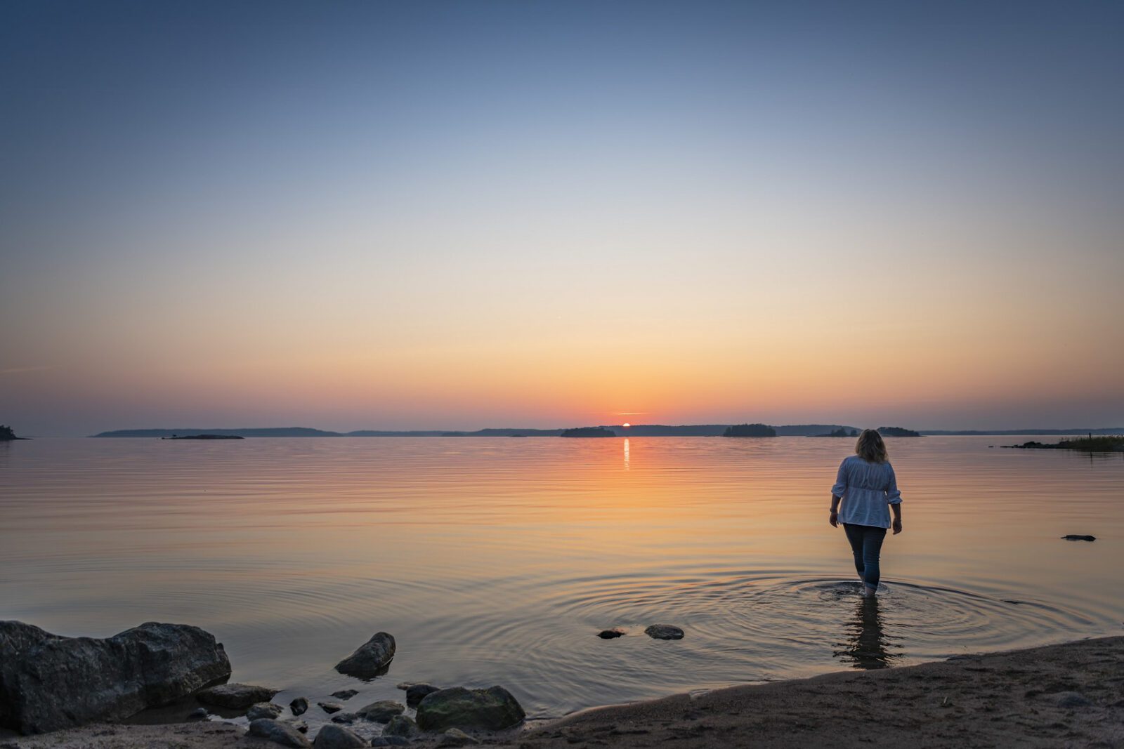 Nainen kävelee merenrannalla katsellen kohti merta ja auringonlaskun värjäämää taivasta. 