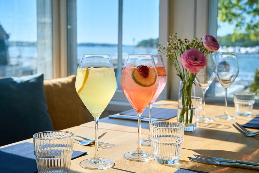 Kesäisen värikkäitä juomia ja leikkokukkia pöydällä, jonka takaa ikkunasta näkyy merimaisema.