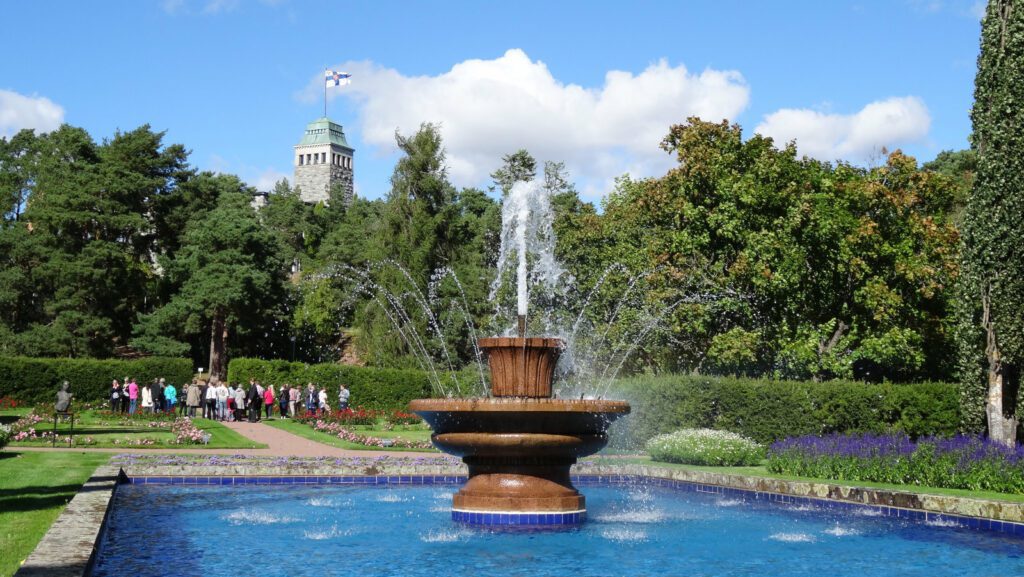 Kultarannan puutarhan iso suihkulähde ja sininen allas, vesi suihkuaa korkealle. Takana Kultarannan kivilinna, jonka tornissa liehuu Suomen lippu.