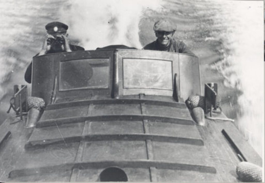 Mustavalkoinen valokuva kahdesta miehestä, jotka ajavat venettä kovaa kyytiä.