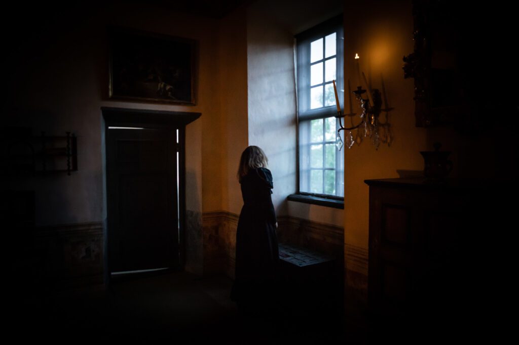 Nainen seisoo hämärässä Louhisaaren kartanolinnan ikkunan edessä katsellen ulos.