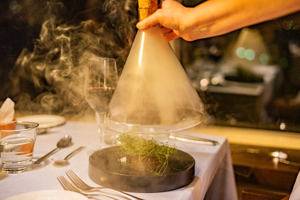 Ravintolan pöydässä tarjoilija nostaa lasisen kartion pois annoksen päältä, kartion alta tulee savua, joka ympäröi lautasella olevan pihviannoksen.
