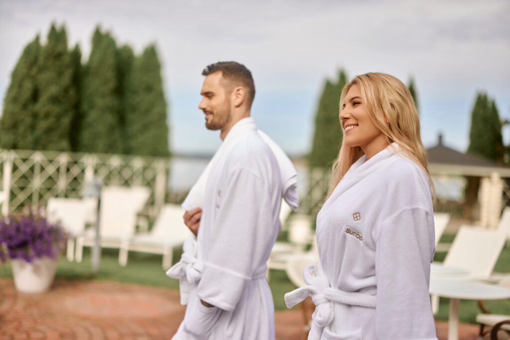 Nainen ja mies kävelevät valkoiset kylpytakit yllään pyyhkeet olalla Naantalin Kylpylän ulkoallasosastolla kesäisenä päivänä.