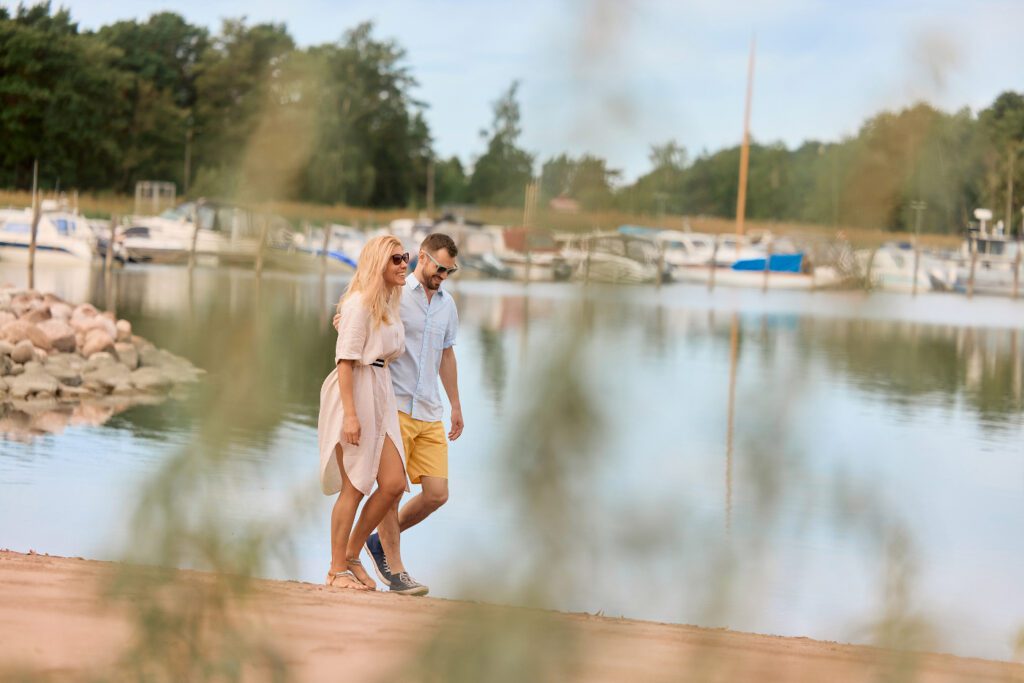 Pariskunta kävelemässä Naantalin Kylpylän hiekkarannalla, taustalla venesatama.